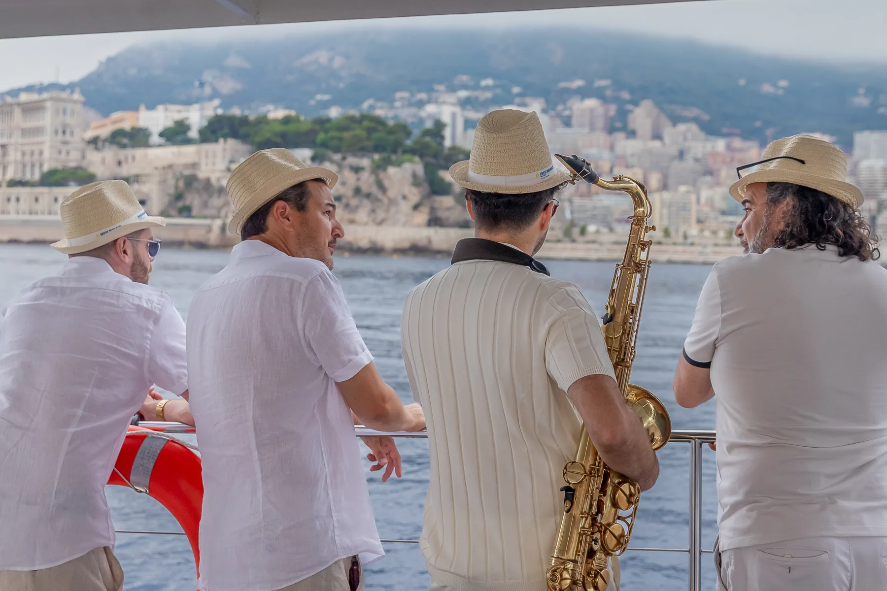 G&auml;ste mit Fayat-Monaco-Strohh&uuml;ten und der Saxofonist an der Reling des Katamarans, gegen&uuml;ber dem Felsen von Monaco